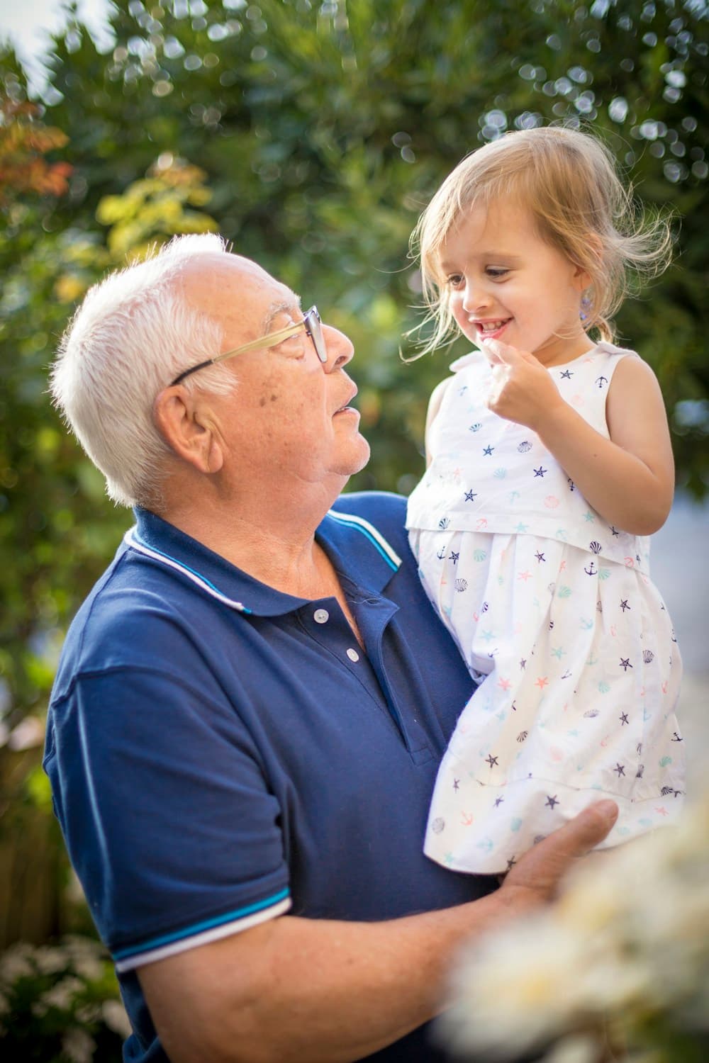 In a garden, a white-haired grandfather with glasses holds a little girl; they look at each other and she smiles.